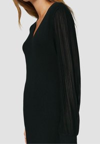 Robe en tricot noir avec décolleté en V et manches longues présentant une texture transparente côtelée. Corps ajusté et manches évasées.
