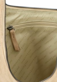 Intérieur d'un sac à main en suède beige avec une poche zippée et une doublure beige clair arborant le nom de la marque en relief, accentué par une fermeture éclair sombre.