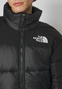 Zbliżenie na osobę noszącą czarną kurtkę puchową The North Face z wysokim kołnierzem i zamkiem błyskawicznym z przodu.
