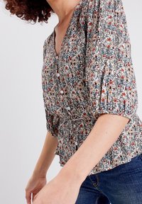 Blusa floreale con scollo a V, maniche corte a sbuffo, chiusura a bottoni frontale e vita arricciata. Il motivo presenta piccoli fiori multicolori su uno sfondo chiaro.