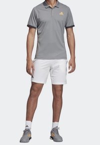 Pánský sportovní outfit skládající se z světle šedého polo trika s černými detaily a oranžovým logem, doplněný o bílé šortky a šedé sportovní boty.