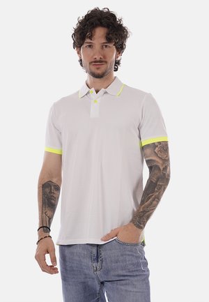 Uomo con capelli ricci che indossa una polo bianca con bordatura gialla e jeans blu, mostrando tatuaggi dettagliati sulle maniche di entrambe le braccia.