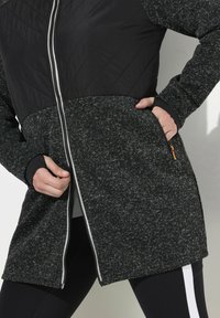 Langer Mantel aus strukturiertem schwarz-grauen Stoff, mit einem gesteppte oberen Teil, Frontreißverschluss und Taschen mit orangefarbenen Akzenten.
