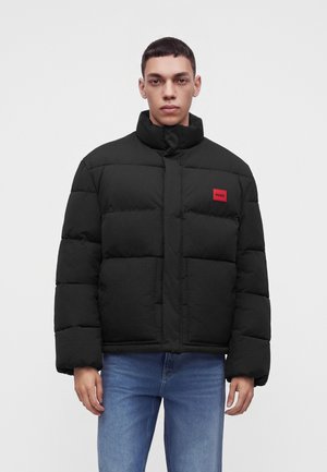 Homme portant une doudoune noire avec un patch rouge Hugo sur la poitrine et un jean bleu, debout devant un fond blanc uni.