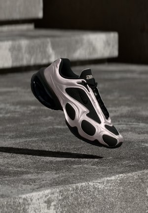 Sporty sort og sølvfarvet sneaker med mesh- og glatte teksturer, svævende over betontrapper i en udendørs bymæssig miljø.