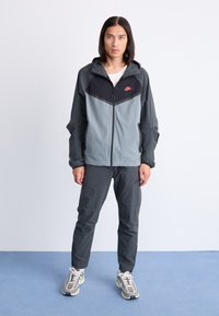 Men's athletic track jacket and pants in grey and black, featuring a zip closure, hood, and contrasting panel design with a small logo.