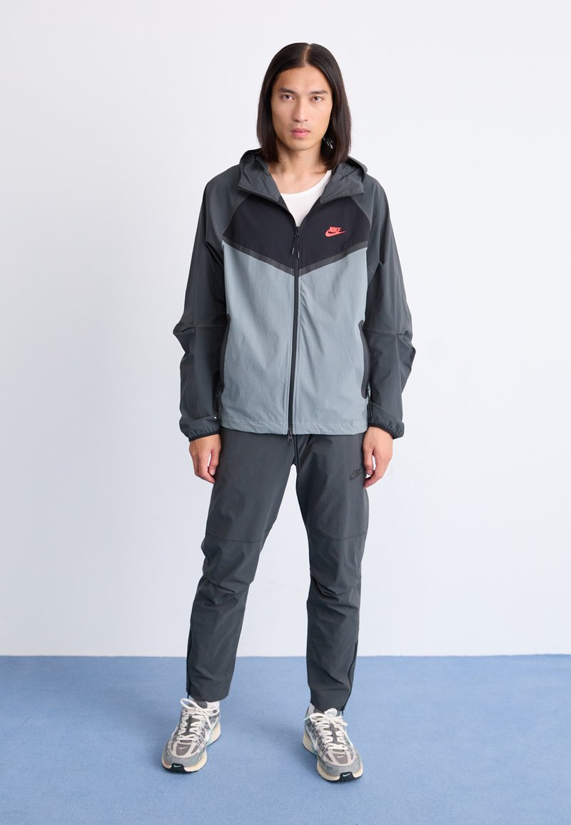 Męska sportowa kurtka na bieganie i spodnie w kolorze szarym i czarnym, z zamkiem błyskawicznym, kapturem oraz kontrastowym wzorem paneli z małym logo.