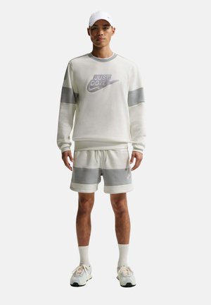 Hombre con sudadera y pantalones cortos Nike blancos y grises, gorra blanca, calcetines y zapatillas, de pie sobre un fondo liso.