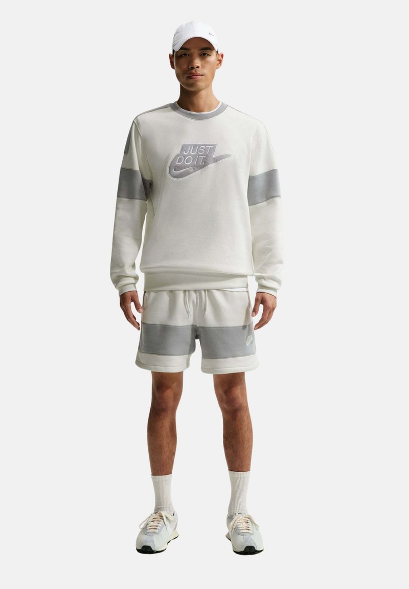 Hombre con sudadera y pantalones cortos Nike blancos y grises, gorra blanca, calcetines y zapatillas, de pie sobre un fondo liso.
