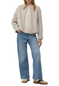 Beige bomberjack met een rits aan de voorkant, gecombineerd met een crèmekleurig overhemd met knopen en lichtblauwe wijde jeans. Tan en witte sneakers maken de outfit compleet.