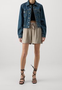 Veste en jean bleu foncé, crop top noir, short plissé beige et sandales à talons noires avec lanières, semelle plate et détails noués.