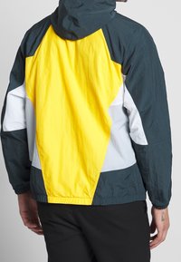 Color-blocked jacket featuring yellow, dark green, gray, and white panels. Lightweight fabric with a hood and elastic hem and cuffs.