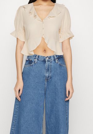 Kvinde iført en beige cropped bluse med flæser og knapper kombineret med højtaljede løse blå denimjeans.