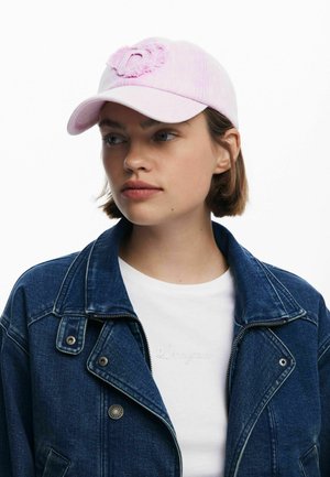 Gorra de béisbol rosa claro con una letra "D" bordada texturizada, llevada con una chaqueta de mezclilla y una camiseta blanca con letras sutiles.