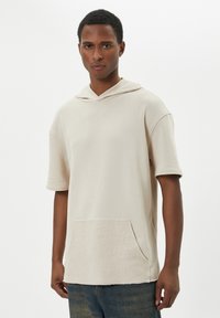 Beige huppari, jossa on lyhyet hihat, etutasku ja rakennehelma. Liukas kangas, jossa on rento istuvuus.