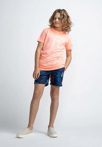 Camiseta rosa claro con texto blanco, combinada con pantalones cortos azul marino con un estampado floral. La modelo lleva zapatos beige sin cordones.