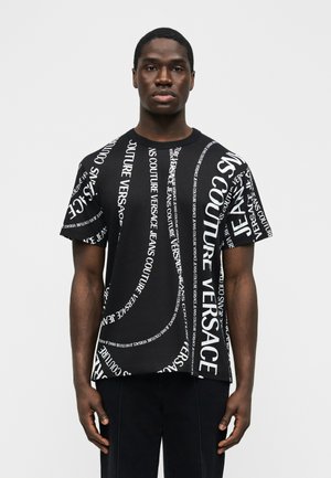 Νεαρός άνδρας που φοράει μαύρο T-shirt με λευκά διαγώνια μοτίβα κειμένου "Versace Jeans Couture", στέκεται μπροστά σε ένα απλό φωτεινό φόντο.