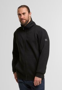 Mann mit Bart, der eine schwarze, strukturiert zip-feste Jacke mit hohem Kragen und dunkelblauen Hosen trägt, steht vor einem einfarbigen, hellen Hintergrund.