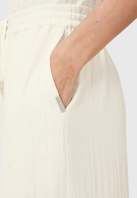 Witte geplooide kleding met een elastische tailleband. Heeft een zijzak met een klein label en een gladde, lichte textuur.
