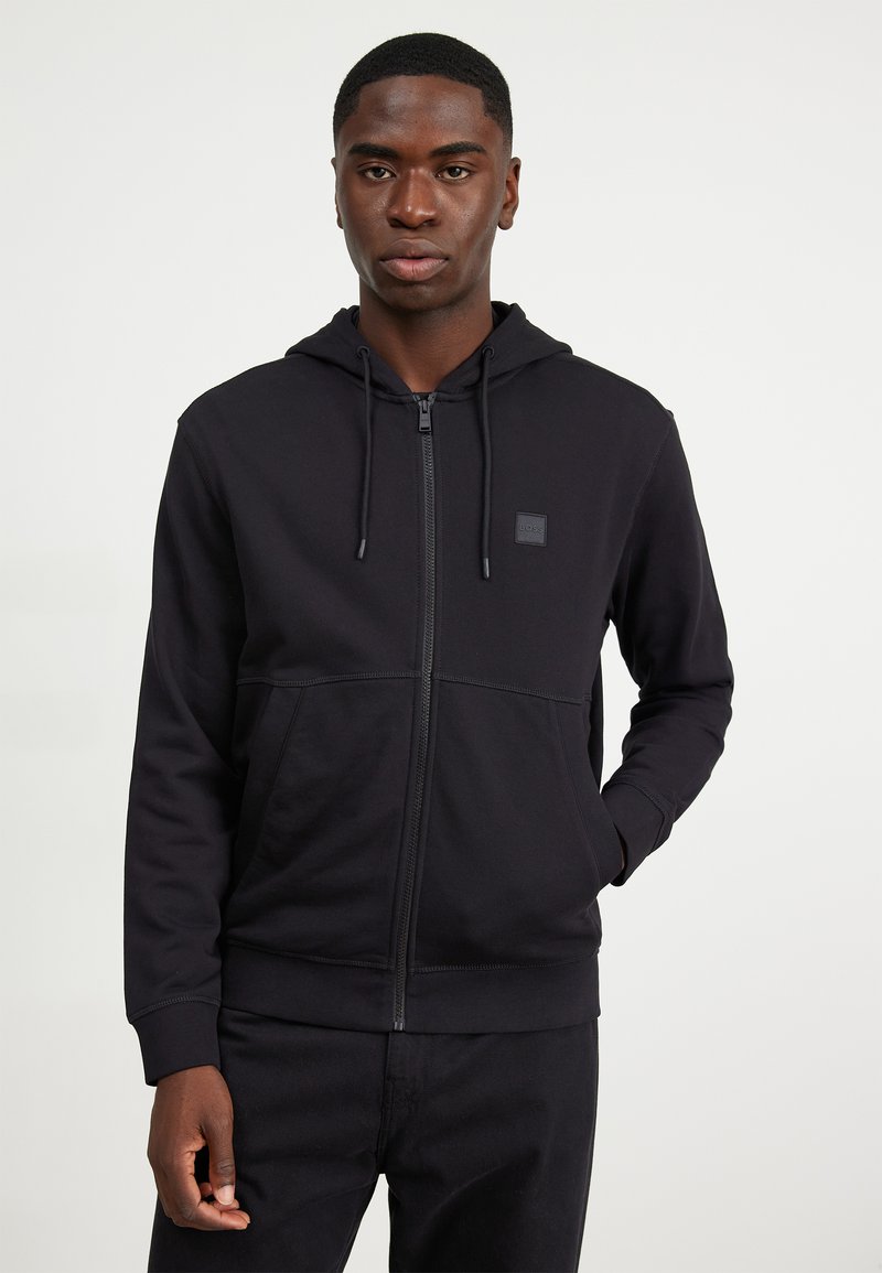 Svart zip-up hoodie i mjukt tyg, med huva och dubbla fickor fram. Inkluderar en liten logopatch på bröstet.