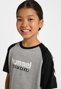 Szara koszulka z czarnymi rękawami, z białym logo "hummel". Krótkie rękawy, luźny krój, okrągły dekolt i kontrastowe akcenty.