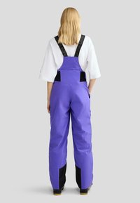 Lila wasserdichte Latzhose mit verstellbaren schwarzen Trägern, Seitentaschen und schwarzen Akzenten an den Knöcheln. Glatte Textur, funktionales Design.