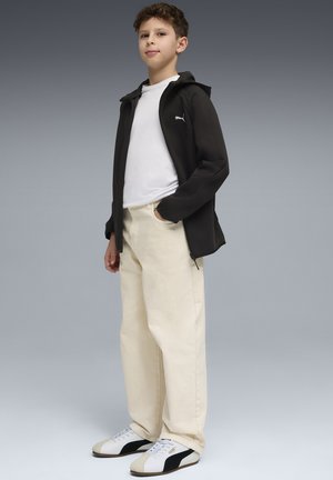 Jongen staat met één hand in de zak, draagt een zwarte hoodie met rits, een wit T-shirt, beige broek en witte en zwarte sneakers.