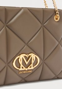 Καφέ καπιτονέ τσάντα με χρυσό λογότυπο "M", αλυσίδα λουρί ώμου και απαλό υλικό. Διακοσμημένη με ραφές σε σχήμα ρόμβου.