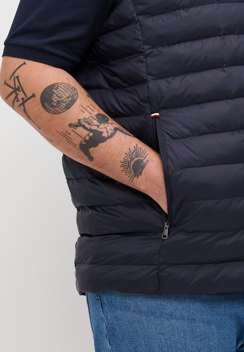 Gilet rembourré bleu marine avec un design matelassé, poche latérale zippée, et un accent contrasté rouge et blanc au bas. Tatouages visibles sur les bras.