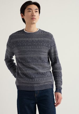 Man met marineblauw gebreide trui met patroon en donkerblauwe spijkerbroek, staand met één hand achter zijn rug, licht omhoog kijkend.