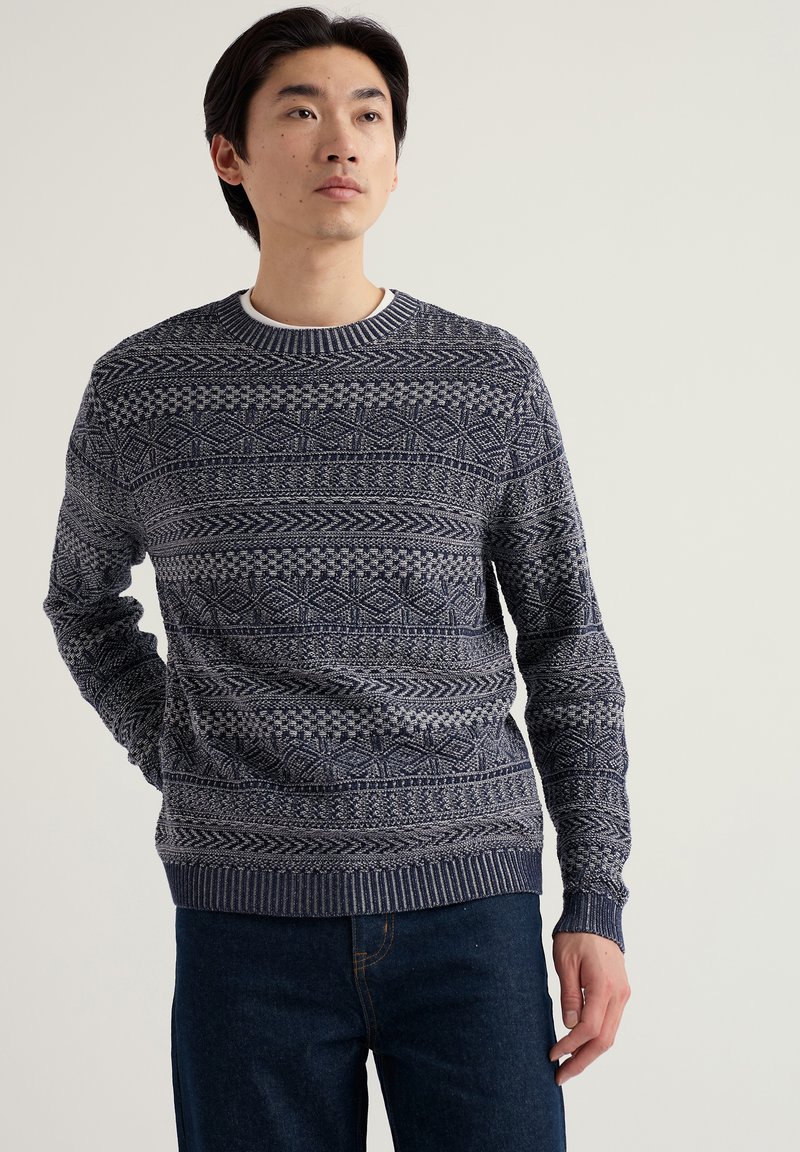 Homme portant un pull en maille bleu marine à motifs et un jean bleu foncé, debout avec une main derrière le dos, regardant légèrement vers le haut.