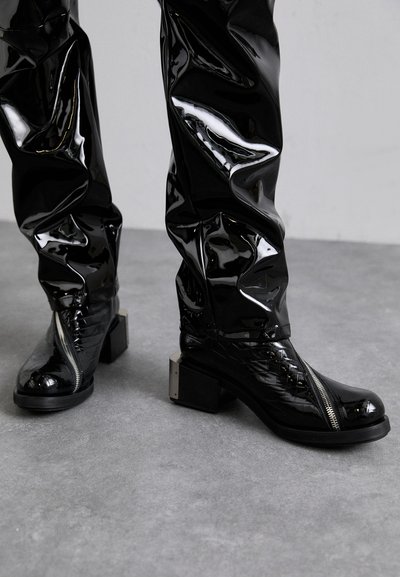 Black patent leather ankle boots with a chunky heel, zip closure, and textured detailing on the sides. Smooth, glossy finish throughout.