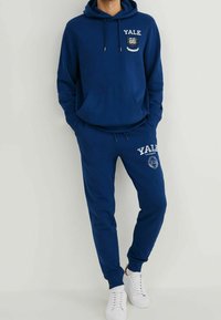 Navy blue hoodie and sweatpants set featuring a front pocket, Yale logo, drawstrings, and ribbed cuffs, complemented by white sneakers.