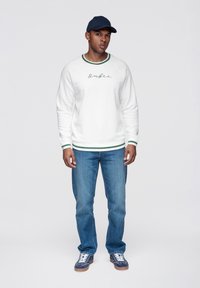Vitt sweatshirt med gröna randiga muddar och halsringning, med texten "Onfire". Matchat med blå jeans och marinblå keps. Vita sneakers med blå detaljer.
