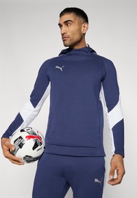 Navyblauwe sweatshirt met witte accentpanelen, capuchon en een buidelzak. Model houdt een voetbal vast met zwarte en rode details. Zilveren logo op de borst.