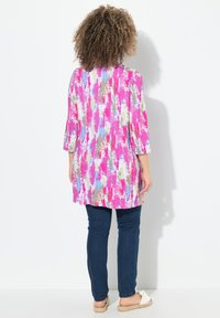 Tunic lunga di un brillante rosa con motivi astratti, caratterizzata da maniche a tre quarti, una vestibilità ampia e spacchi laterali. Abbinata a jeans scuri.