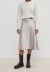 Suéter de punto crema con textura acanalada y detalles laterales, combinado con una falda midi de satén plateado brillante y botas negras por el tobillo.