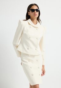 Hvid dobbeltbrystet blazer med runde skulderpuder og matchende tætsiddende nederdel, begge med store knapper; parret med katteøje-solbriller.