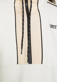 Gros plan sur un sweat à capuche Levi's beige et blanc avec une fermeture éclair noire, des cordons noirs à rayures blanches et des détails de finition noirs.