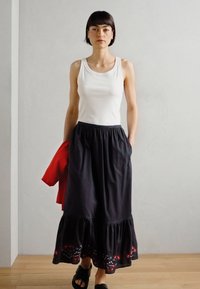 Biely top bez rukávov, čierna maxi sukňa s ruffles na okraji a červenými vyšívanými detailmi, čierne sandále, červená bunda prehodená cez ruku, neutrálny pozadie.
