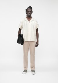 Jaunas vyras dėvi smėlio spalvos kelnes, dryžuotą trumpomis rankovėmis marškinius, pilkus sportbačius, laiko sulankstytą juodą striukę priešais vientisą baltą foną.