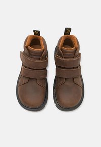 Botas de cuero marrón con dos correas de Velcro, cuello acolchado y suelas de goma negra. Costuras amarillas acentúan el diseño.