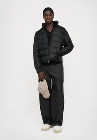 Ung mand i sort pufferjakke, sorte cargo-bukser, hvide sneakers, der holder en beige baseballkasket mod hvid baggrund.
