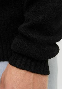 Czarny sweter z dzianiny, rękaw z ściągaczem, strukturalny materiał i luźny krój, zbliżenie na detale materiału i szwów.