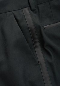 Les pantalons noirs ajustés présentent une texture lisse, une bande satinée élégante sur le côté et une poche latérale à droite.
