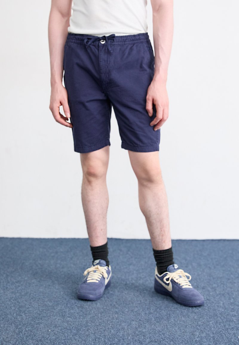Shorts bleu marine en tissu léger, avec une taille élastique et un cordon de serrage, dotés de poches, associés à des baskets bleu marine et crème.