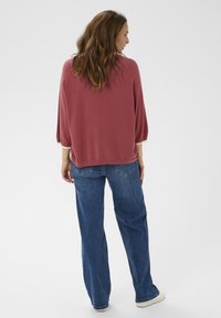 Hosszú barna hajú nő, aki egy laza rózsaszín pulóvert, kék farmert és fehér sportcipőt visel, áll és háttal fordul egy fehér háttér előtt.