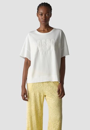 Woman wearing a loose white short-sleeve top with textured lettering and yellow lace wide-leg pants, standing against a plain background.