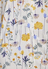Bomullskjorta med knappar som har en vit bakgrund med ett blommönster i gula, lila och gröna nyanser. Noterbar knapppresentation är synlig.