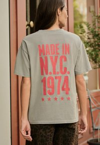 Grå bomulds T-shirt med fed rød tekst "MADE IN N.Y.C. 1974" og fem stjerner. Løs pasform med korte ærmer og rund halsudskæring.